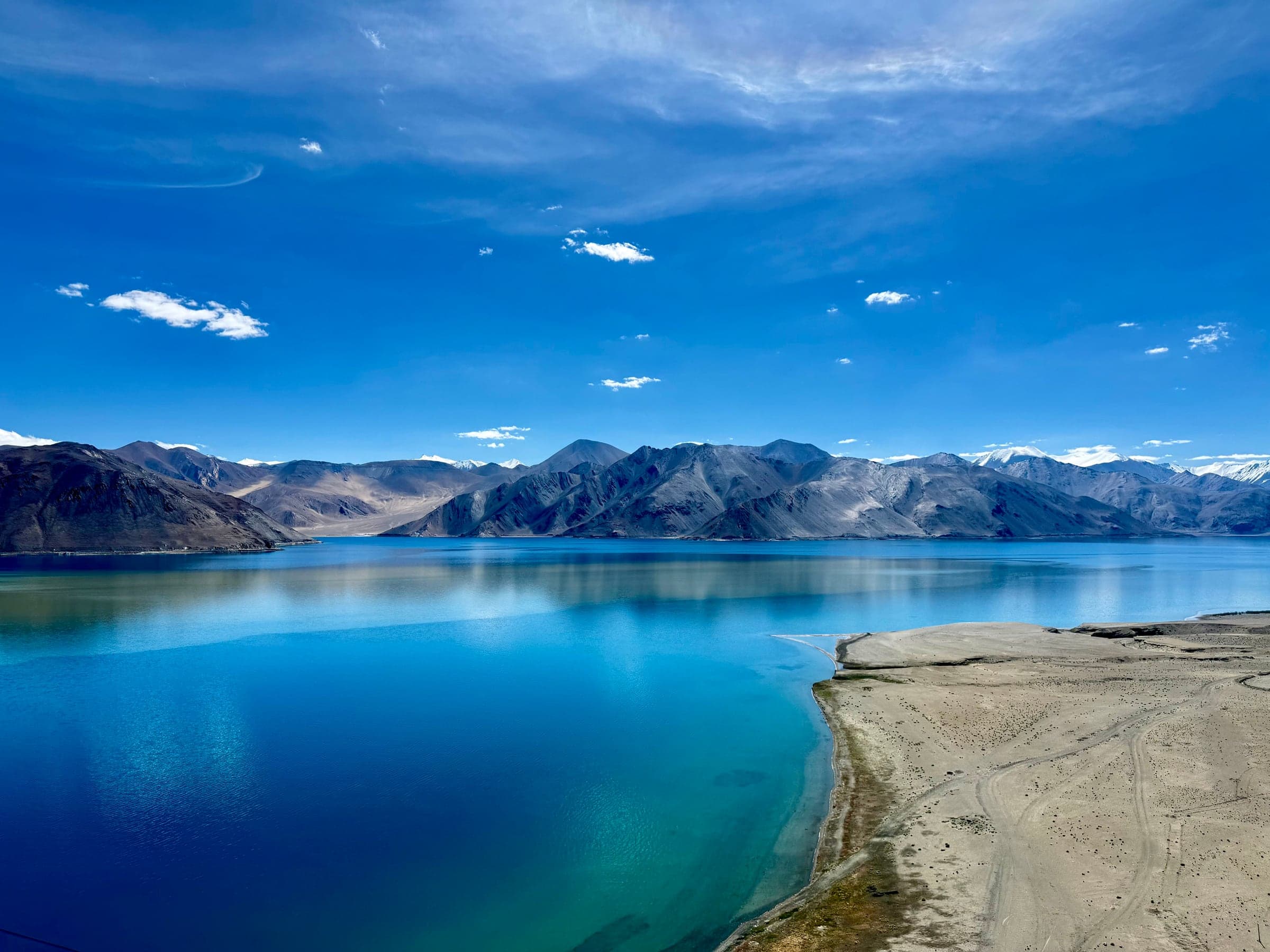 A deep blue high-altitude lake with a sandy spit in the foreground and bare mountains behind