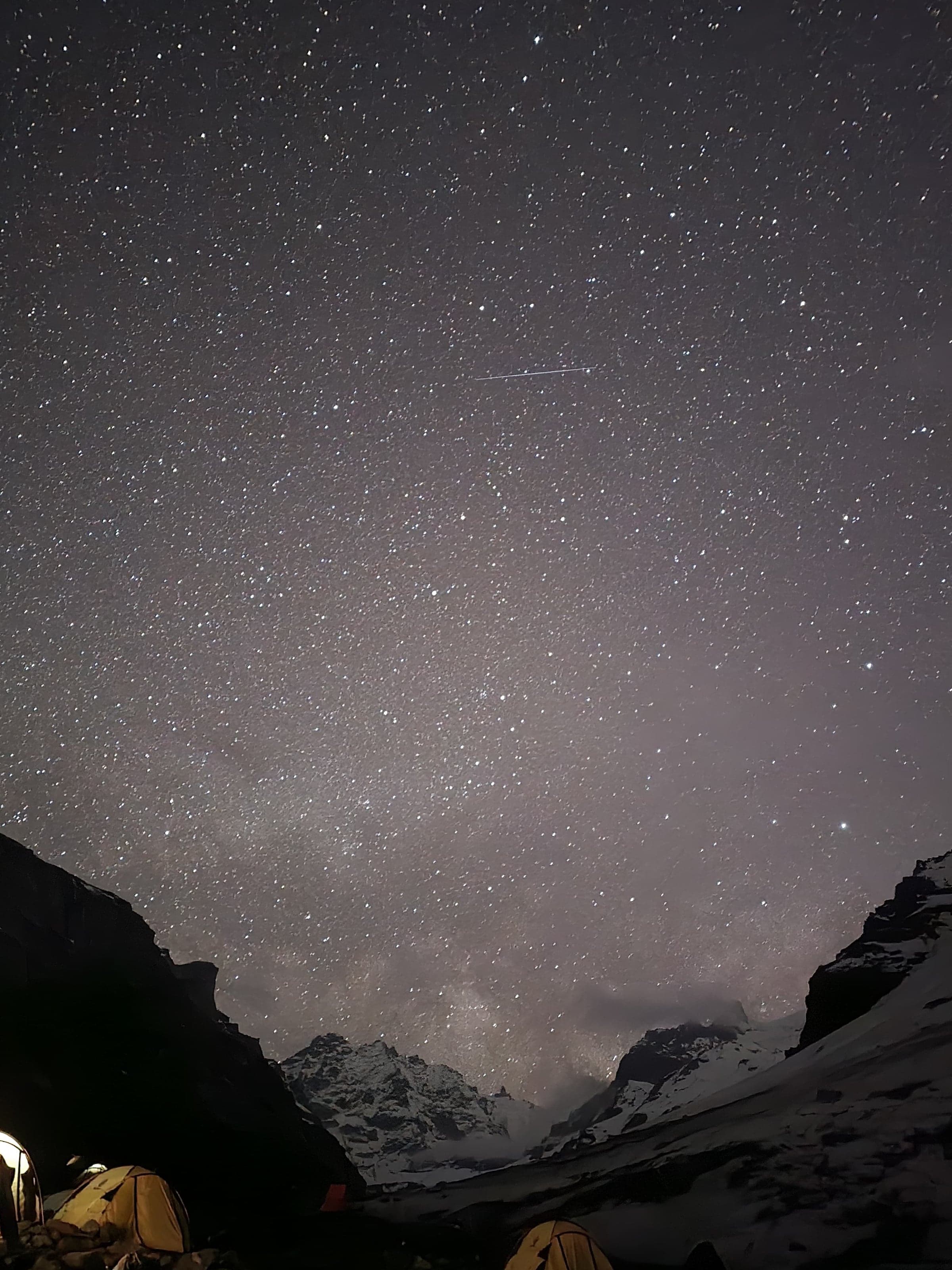 A sky full of stars over a snowy mountain valley, with lit tents at the bottom of the frame