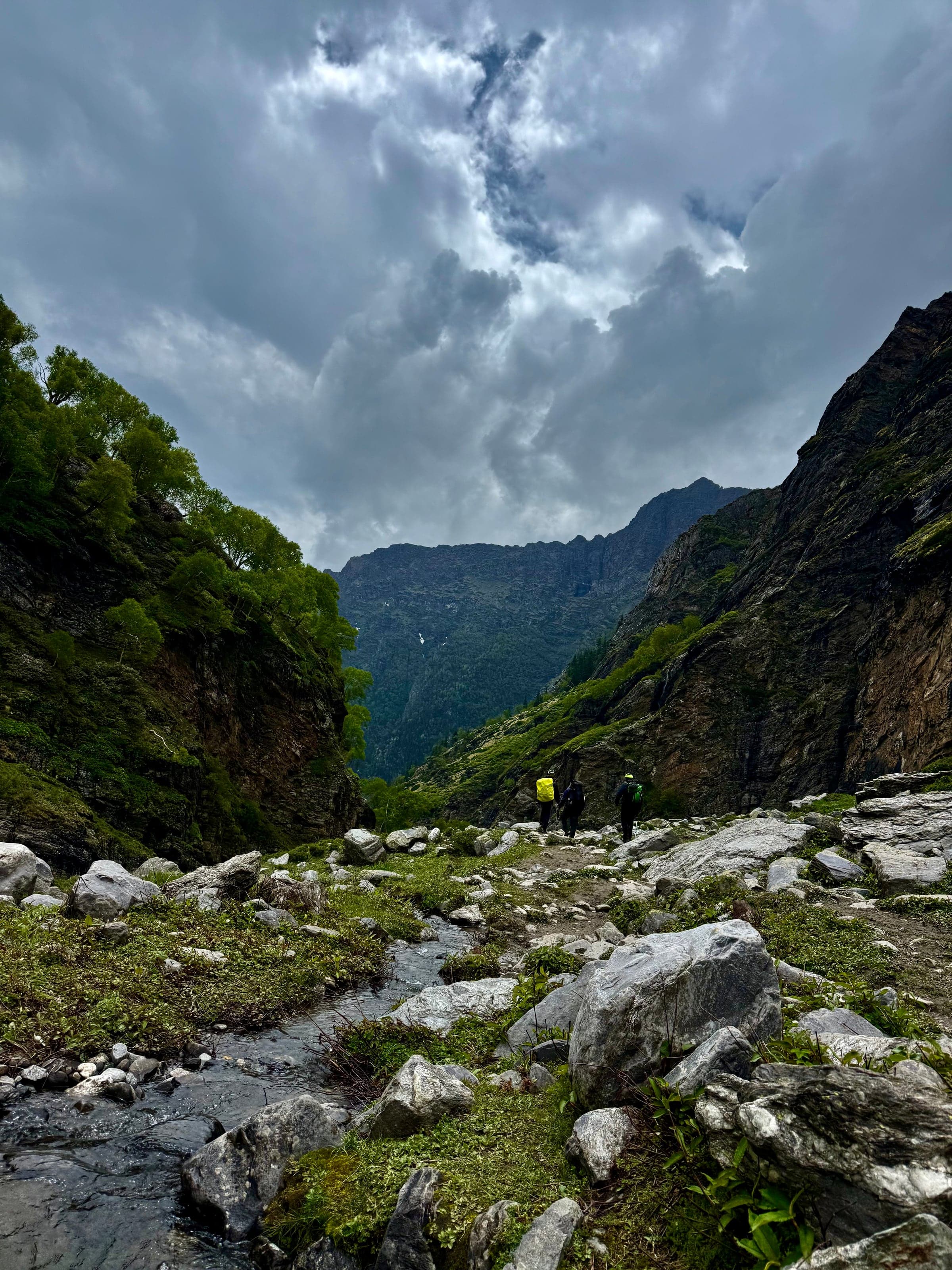 Three trekkers walking down a rocky green valley beside a stream under heavy cloud