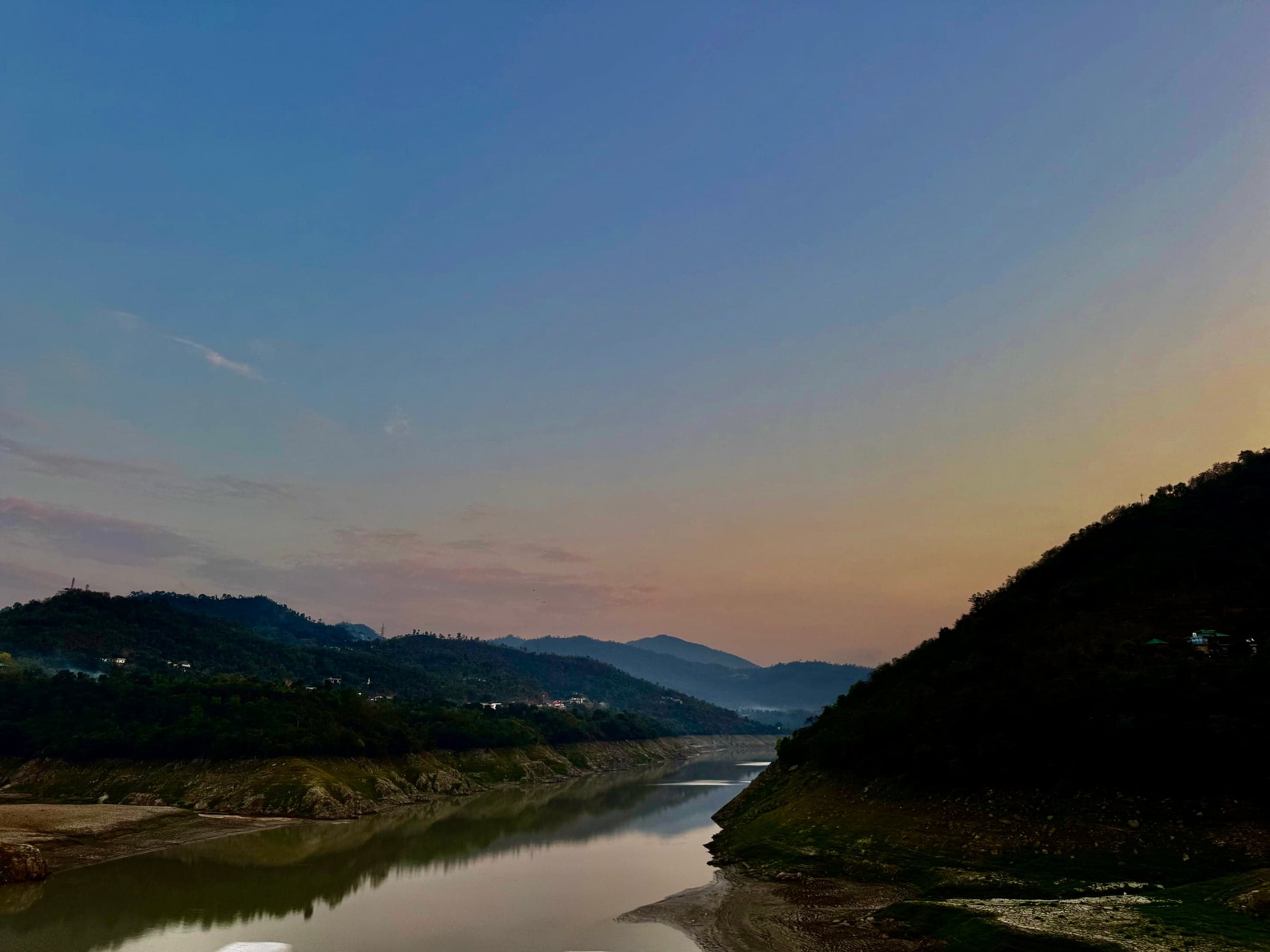 A still river winding between forested hills at dusk, the sky fading from blue to orange