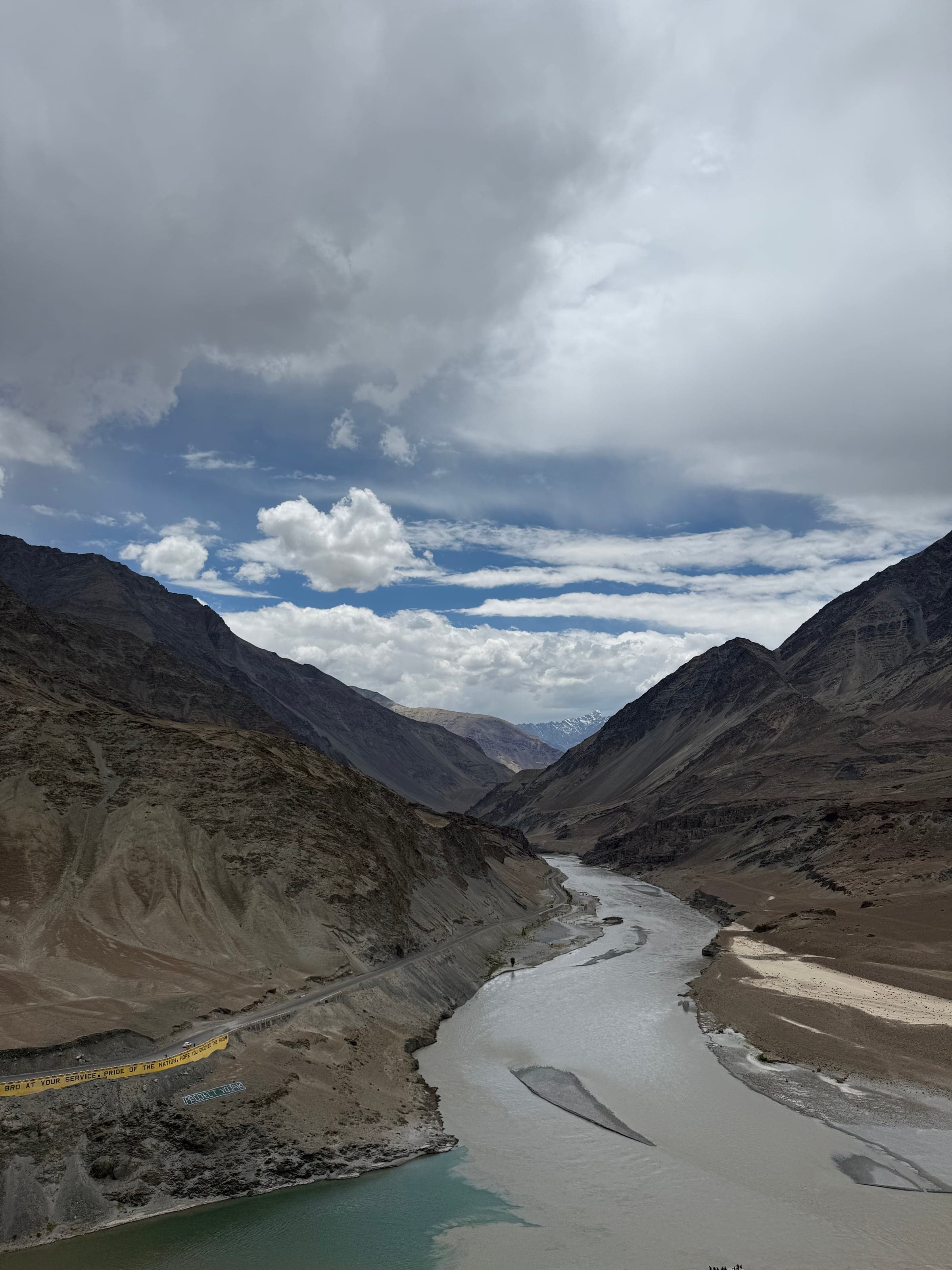 A wide silty river bending through bare brown mountains, with a road along one bank