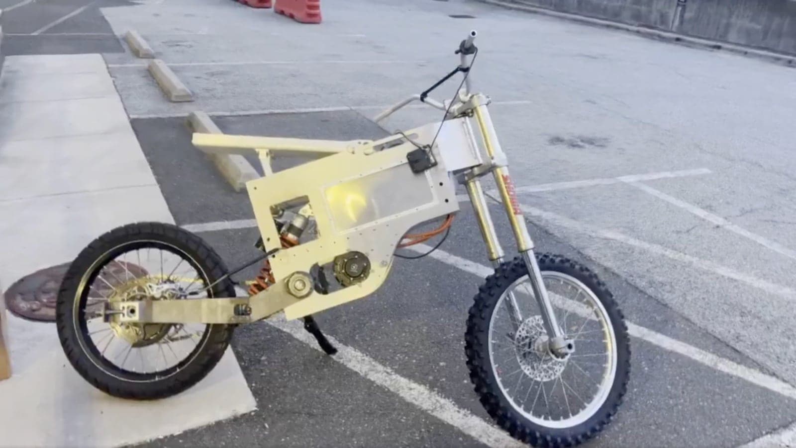 The foldable electric dirt bike standing in a parking lot, bare aluminum frame with the battery bay and rear swingarm visible