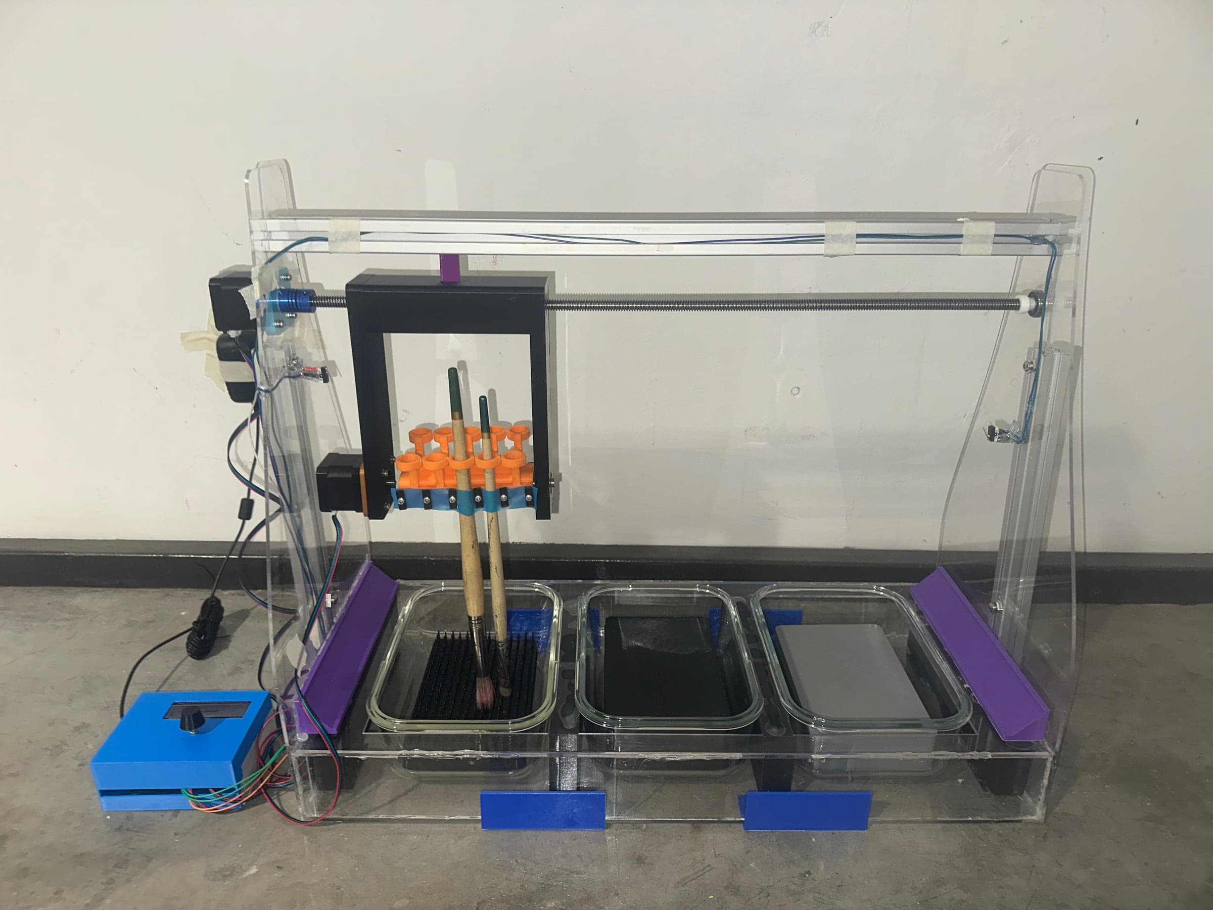 The paintbrush cleaning tool, a clear acrylic frame with a carriage holding two brushes over the first of three glass containers, and a blue control box beside it