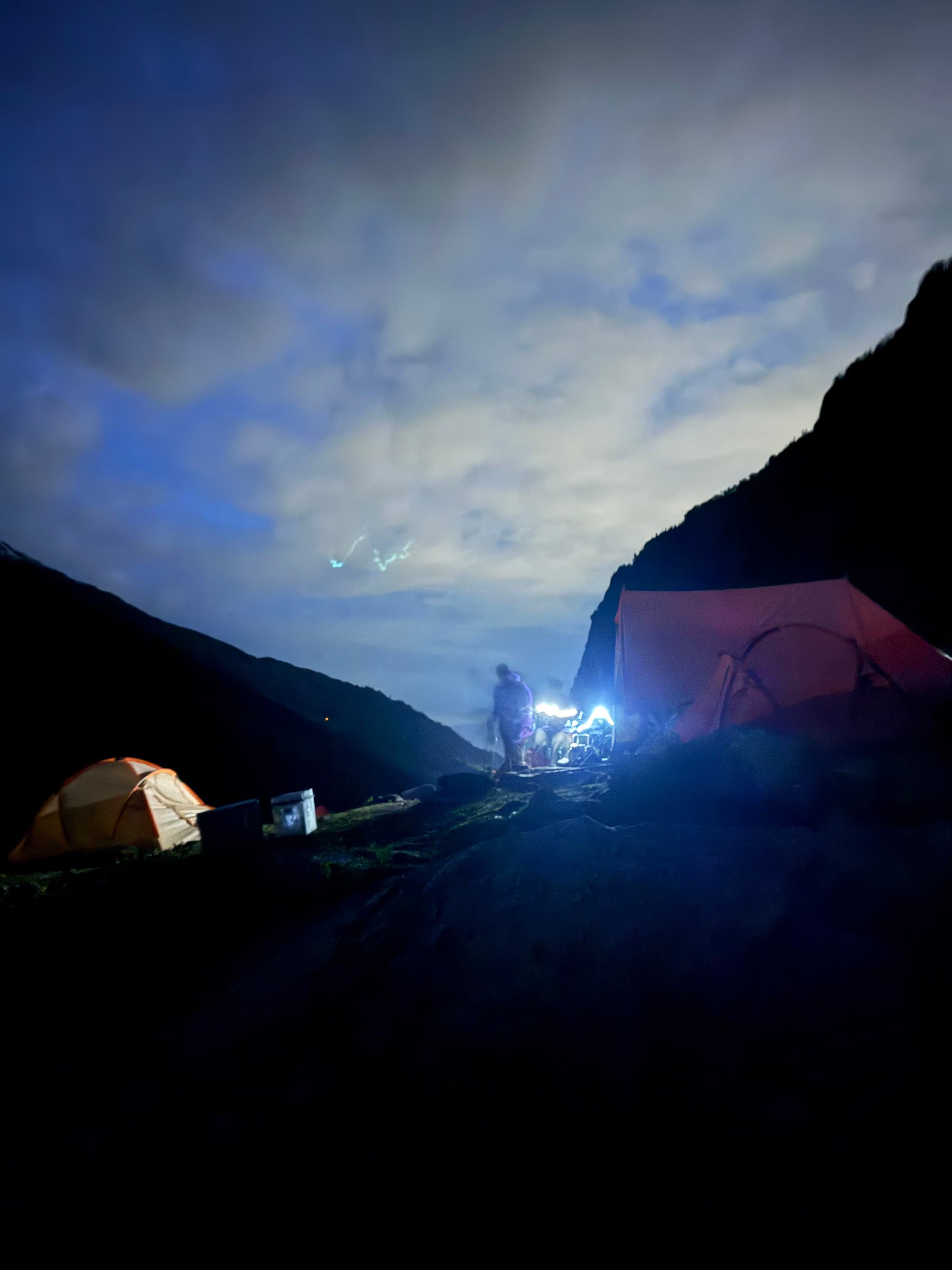 A campsite at night under bright moonlit clouds, with an orange tent and a figure lit by lamps