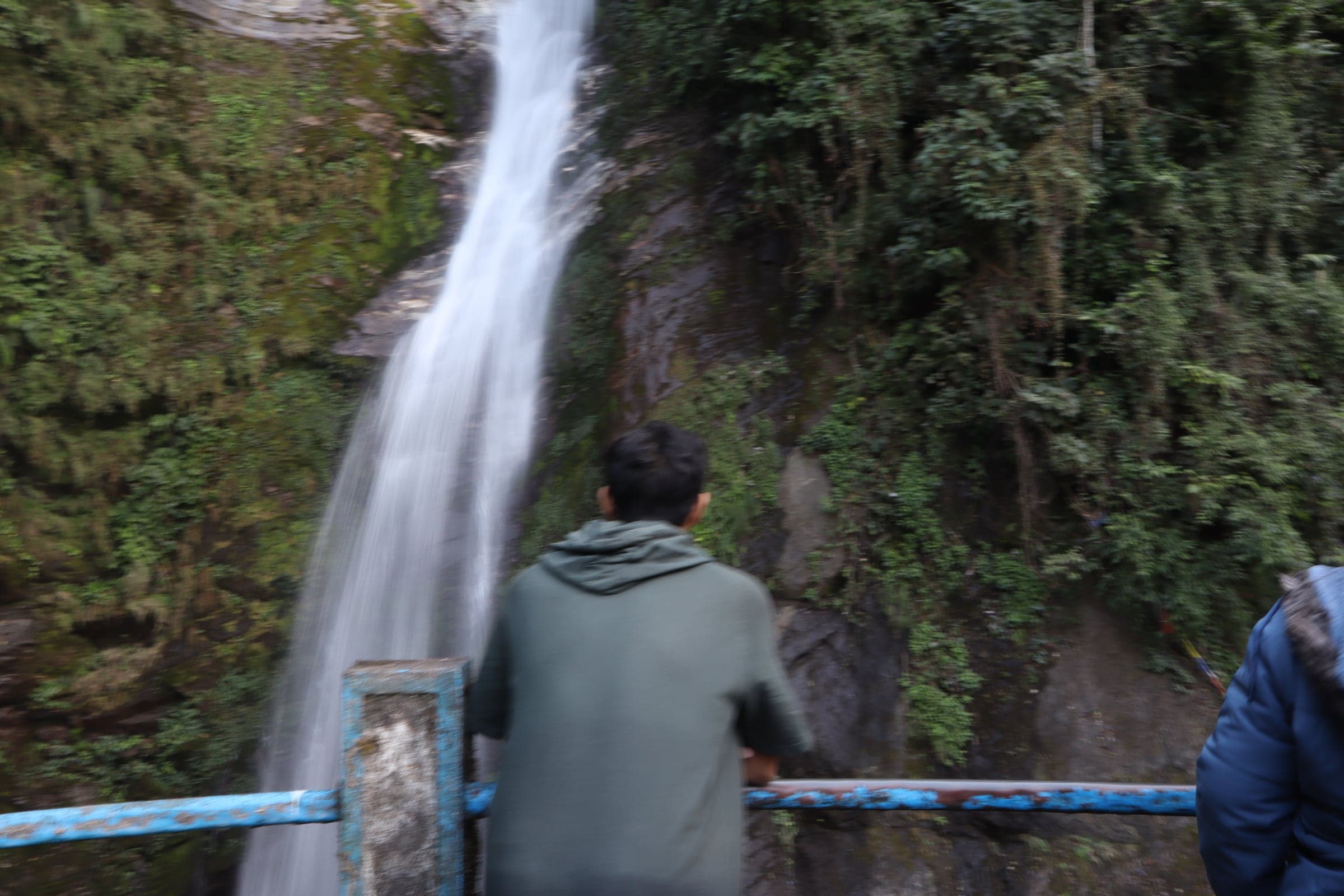 A person in a green hoodie leaning on a railing, watching a tall waterfall